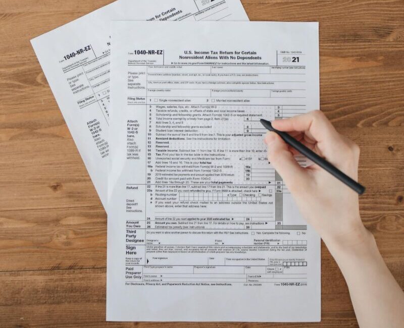 Overhead view of hand writing on tax forms atop a wooden table.