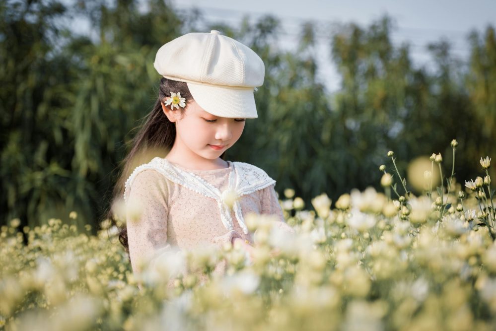 A charming young girl with a cap smiles among blooming flowers in a sunny outdoor setting.