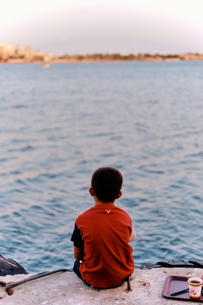 A young boy sits by the sea at sunset in İstanbul, contemplating the horizon.