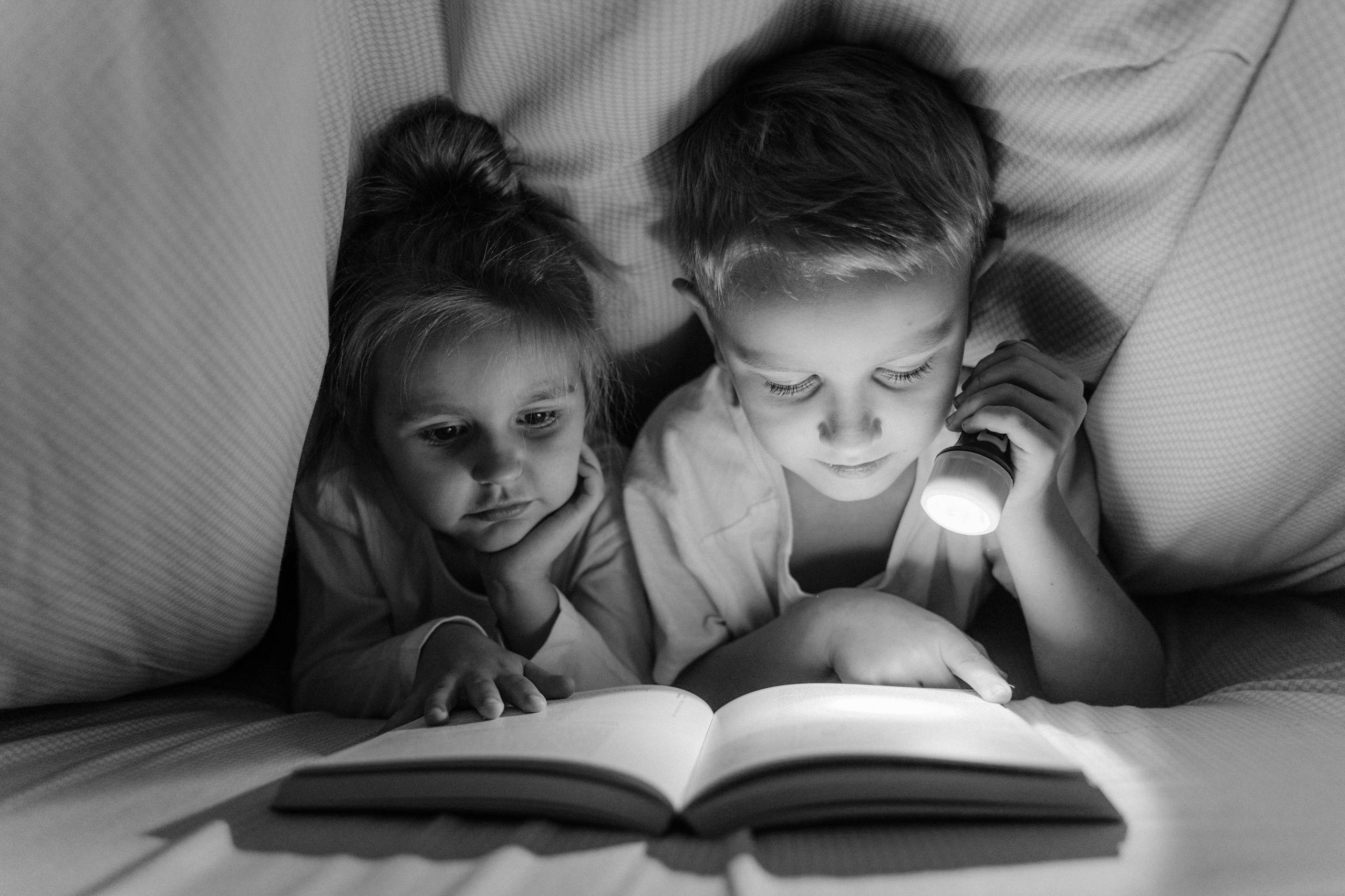 A boy and girl reading with a flashlight under a blanket in grayscale