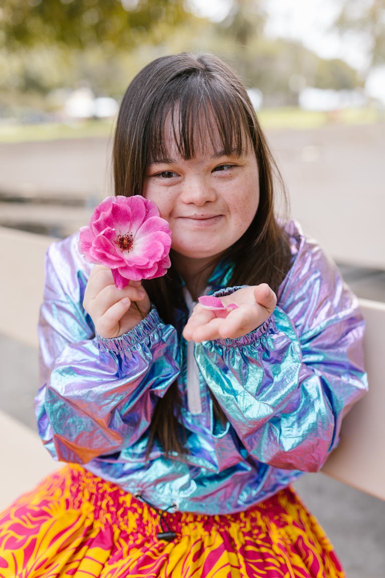 A joyful young woman with long hair holds a pink flower while smiling outdoors, wearing a colorful outfit.