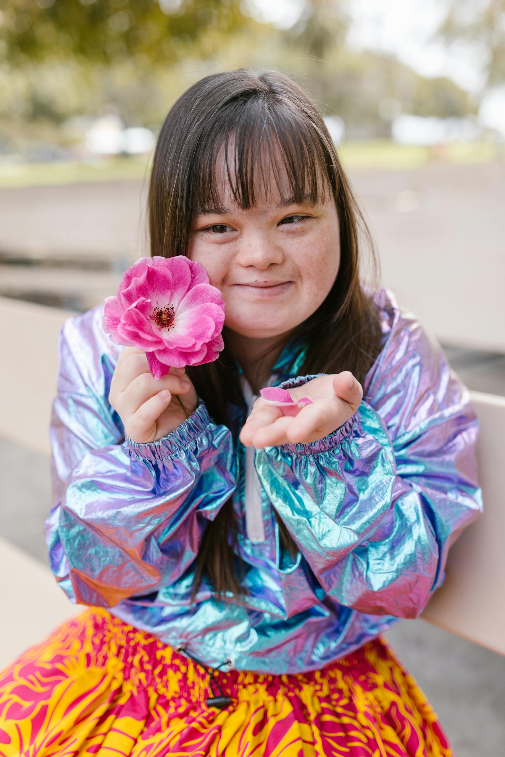 A joyful young woman with long hair holds a pink flower while smiling outdoors, wearing a colorful outfit.