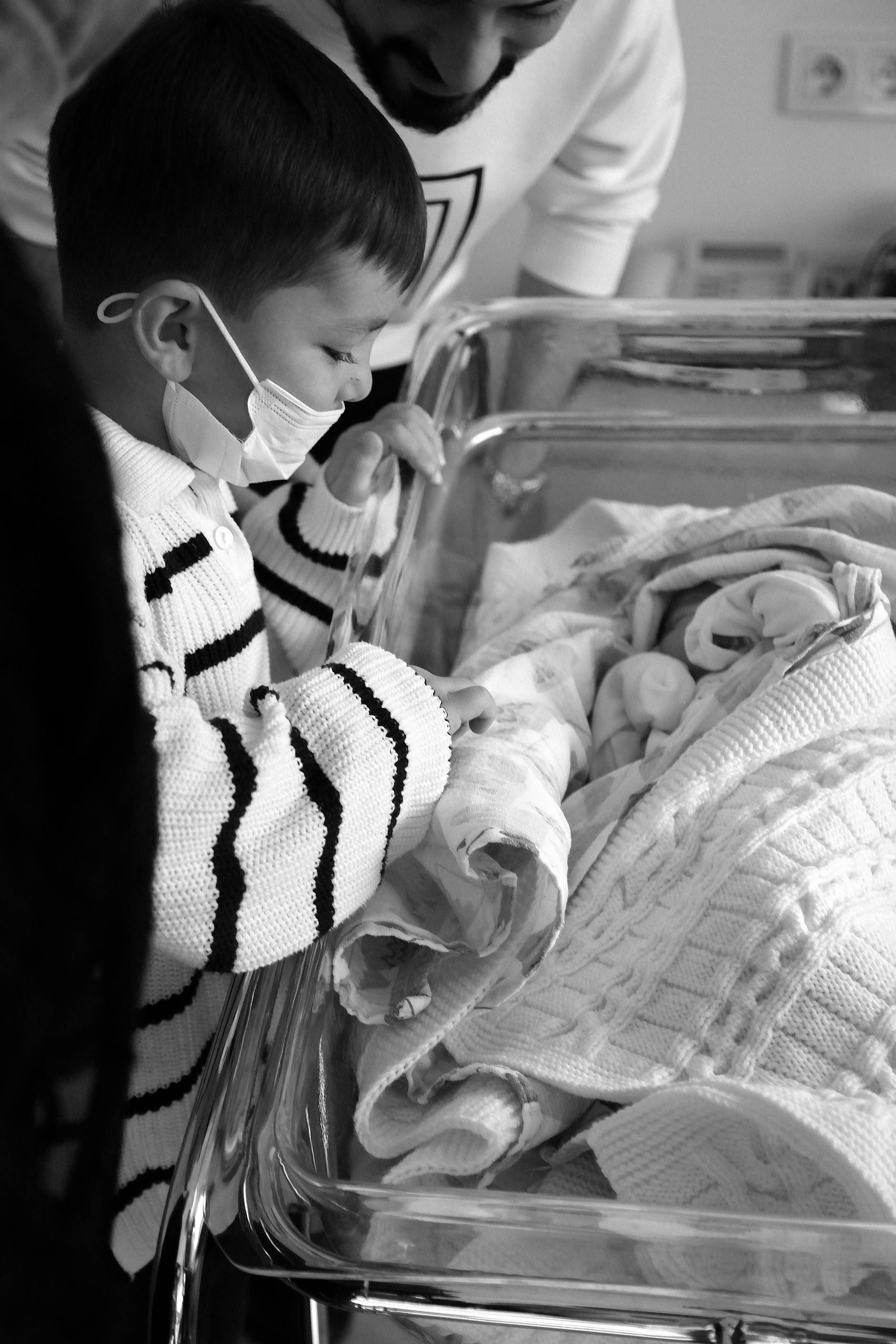 A young child wearing a mask looking at a newborn baby in a hospital setting. Black and white image.