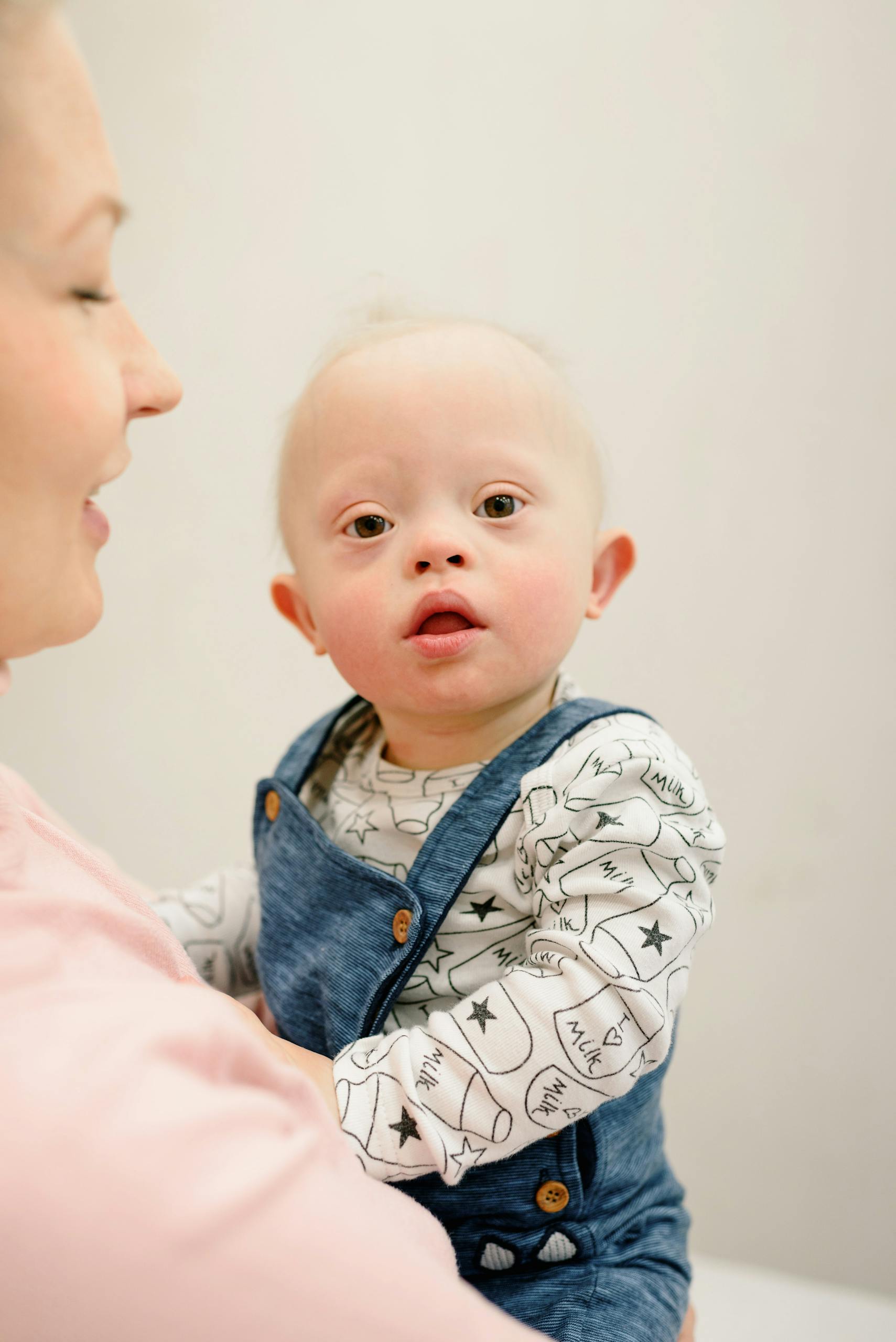 Adorable baby with Down syndrome in blue overalls, held by an adult indoors.