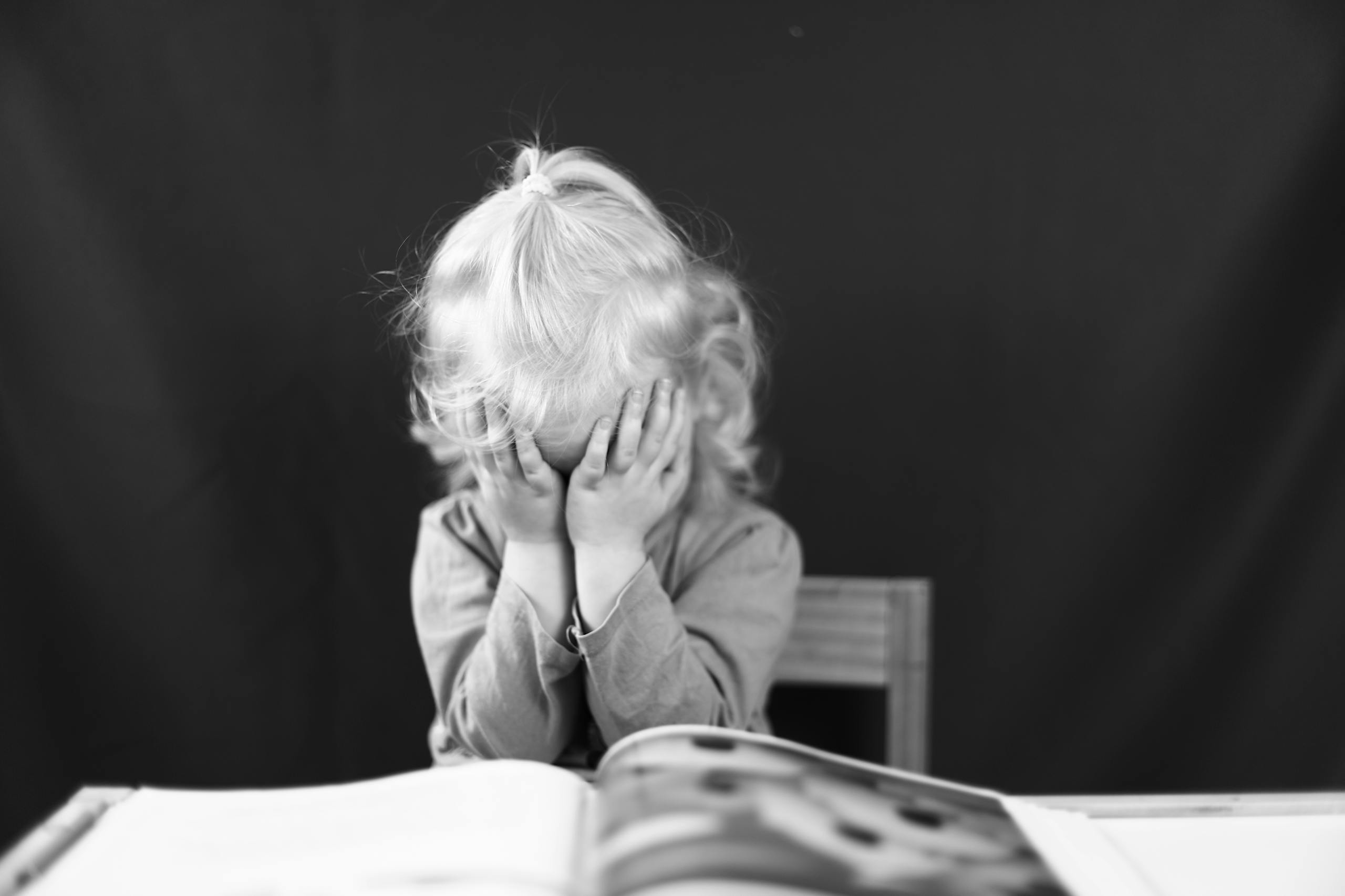 Black and white photo of a young child covering their face with hands, sitting in front of a book.