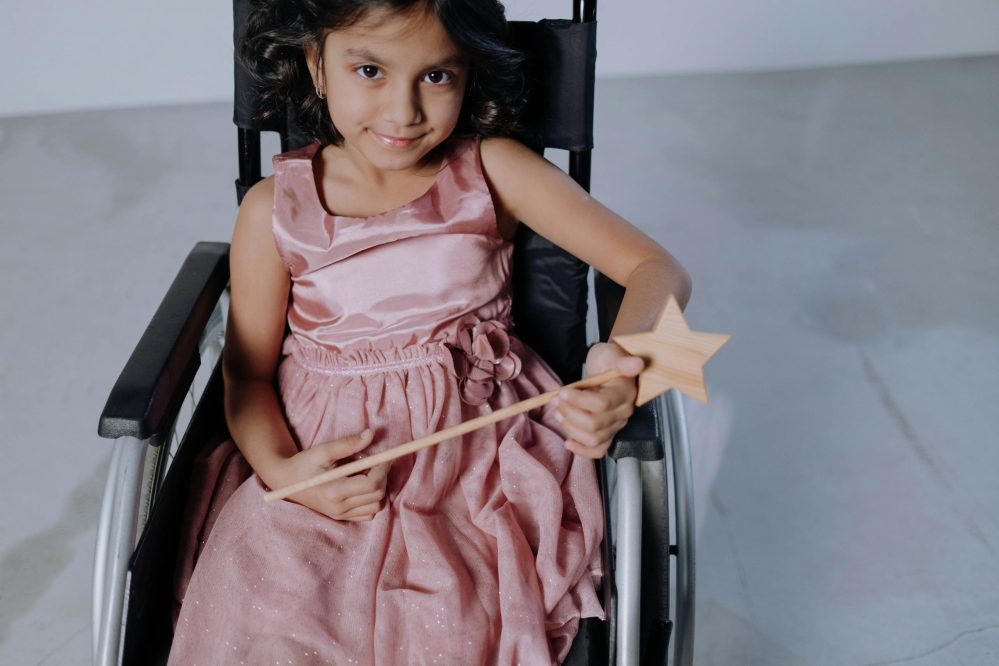Charming young girl in wheelchair joyfully holding a wooden magic wand indoors.
