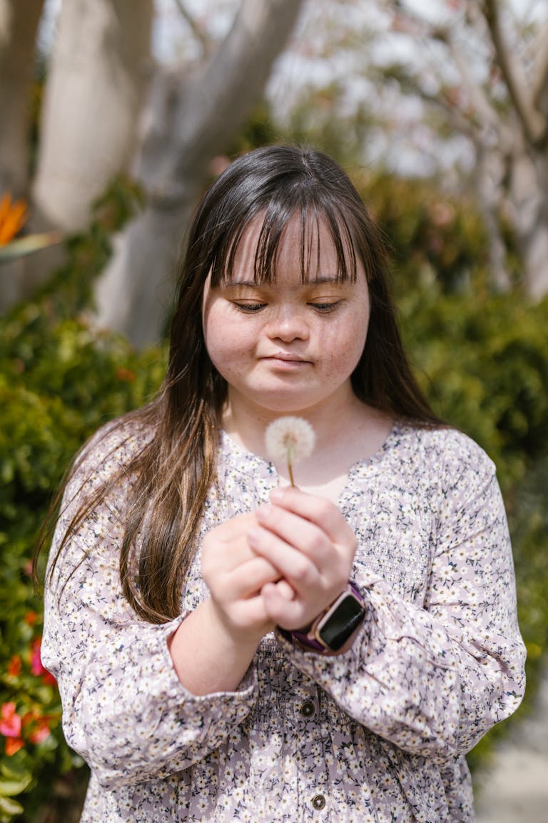 Young girl with Down syndrome holding a dandelion in a garden setting.