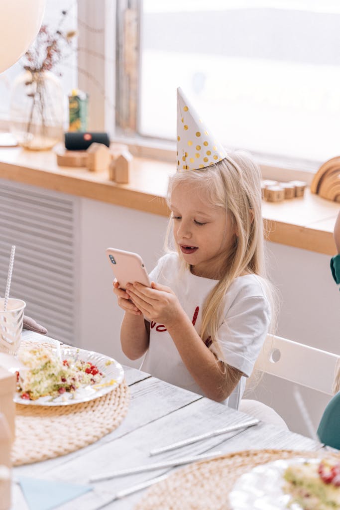 A young girl in a party hat using a smartphone during a virtual party indoors.