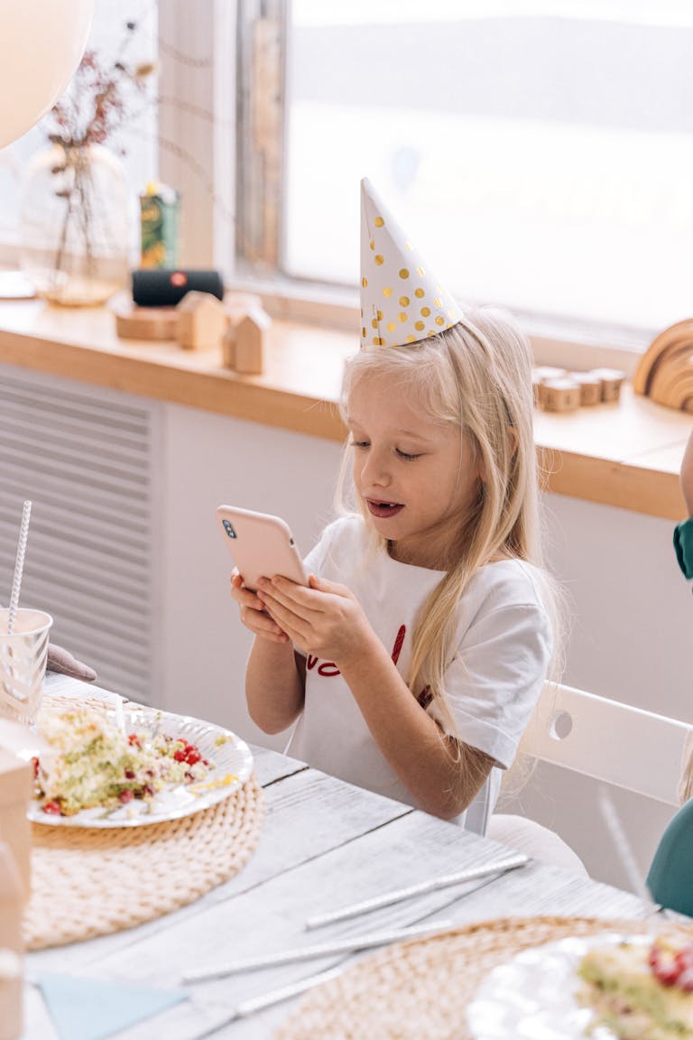 A young girl in a party hat using a smartphone during a virtual party indoors.