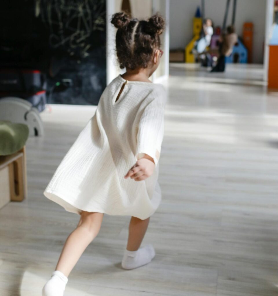 A young girl with hair buns runs joyfully in a sunlit playroom wearing a white dress.