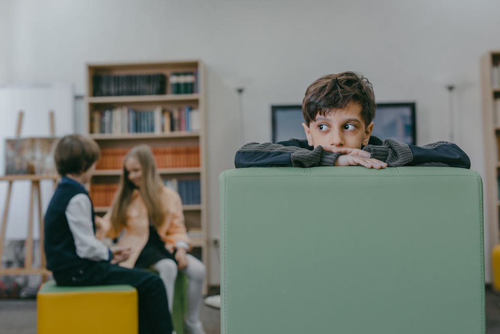 Boy sitting alone on a chair in a classroom while classmates interact in background.