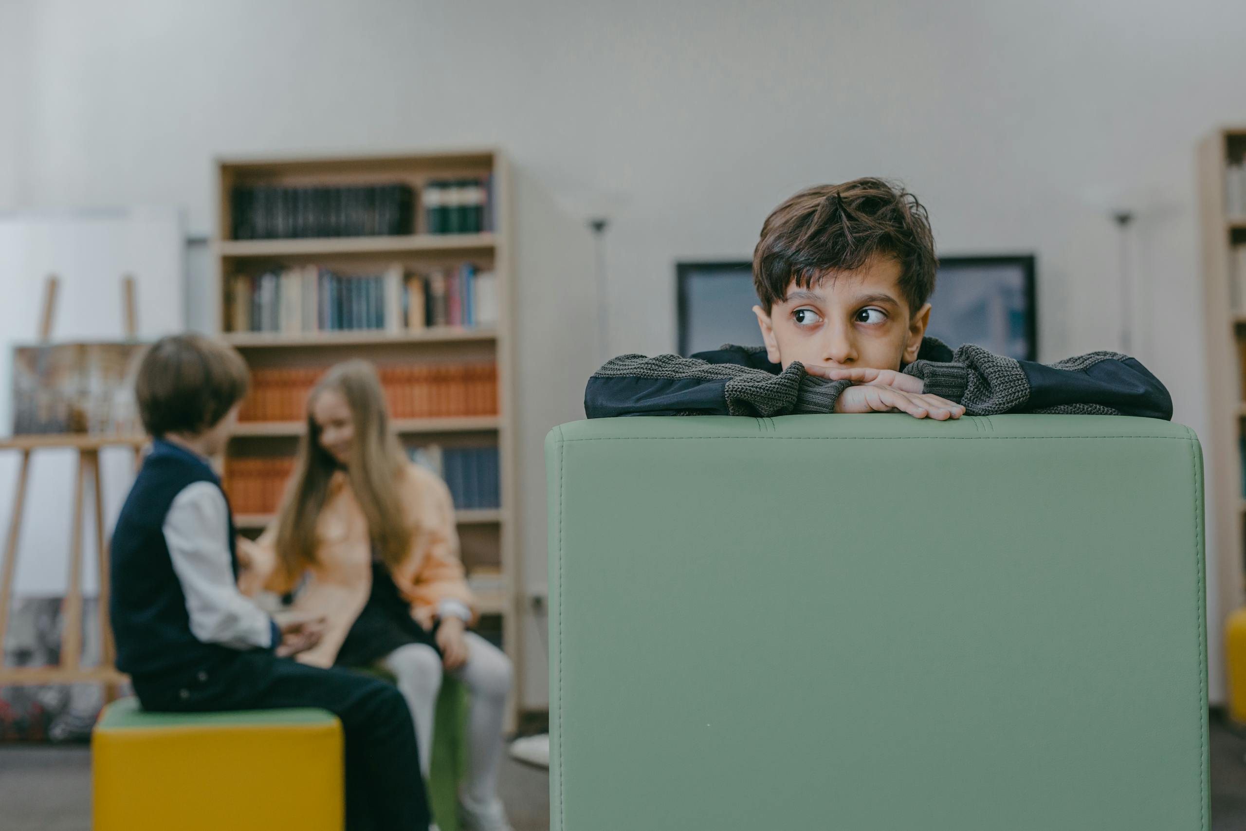 Boy sitting alone on a chair in a classroom while classmates interact in background.