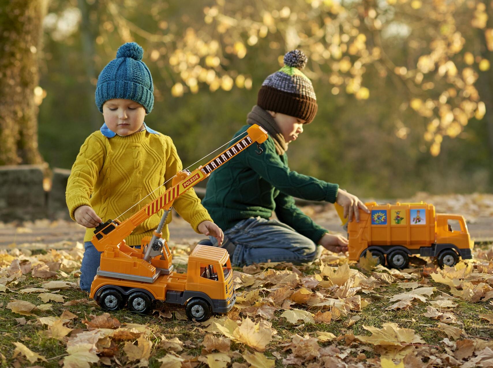 Two kids enjoying autumn outdoors with toy trucks under a tree.